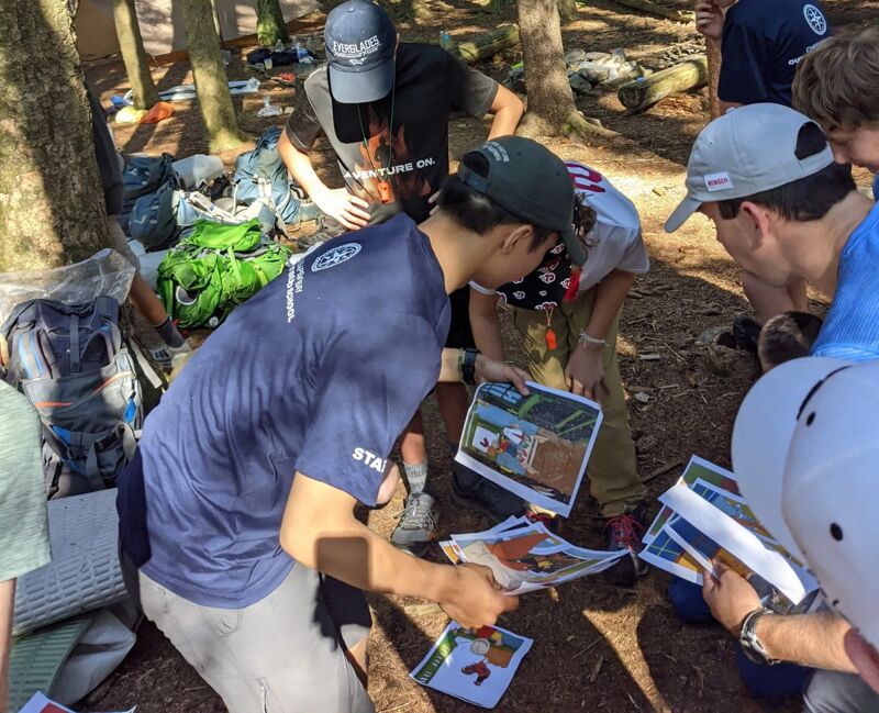 A group of people are gathered in a forest, seemingly engaged in an activity that involves maps or printed materials. They are standing around looking at the items on the ground. The setting is a wooded area with tall trees and dappled sunlight. Some of them are wearing hats. There are backpacks and other gear scattered around, suggesting they might be hiking or camping.